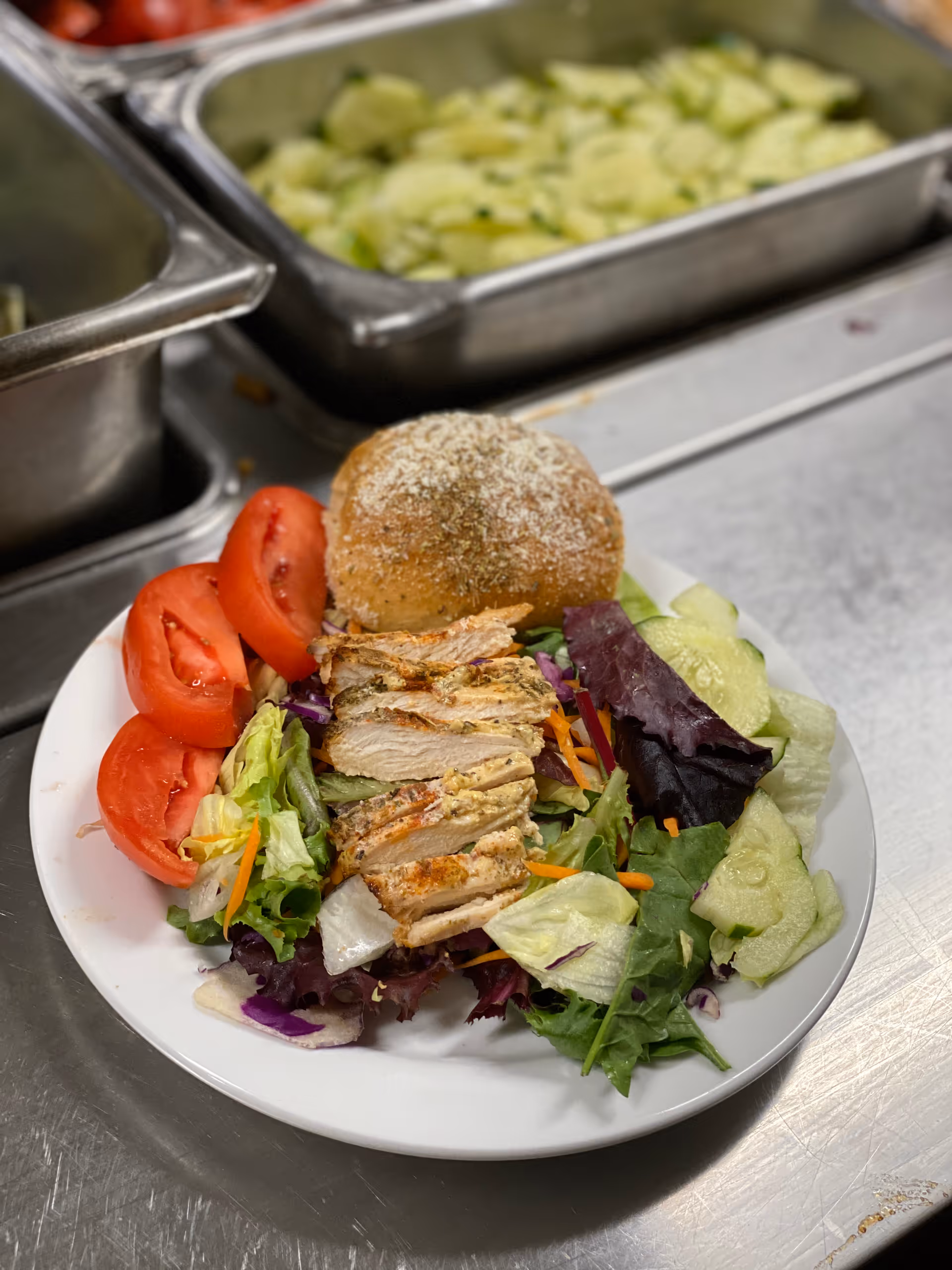 A plate of food featuring grilled sliced chicken breast on a bed of mixed salad greens with sliced tomatoes and cucumbers, accompanied by a seasoned bread roll. The plate is placed on a stainless steel counter with trays of sliced cucumbers in the background.