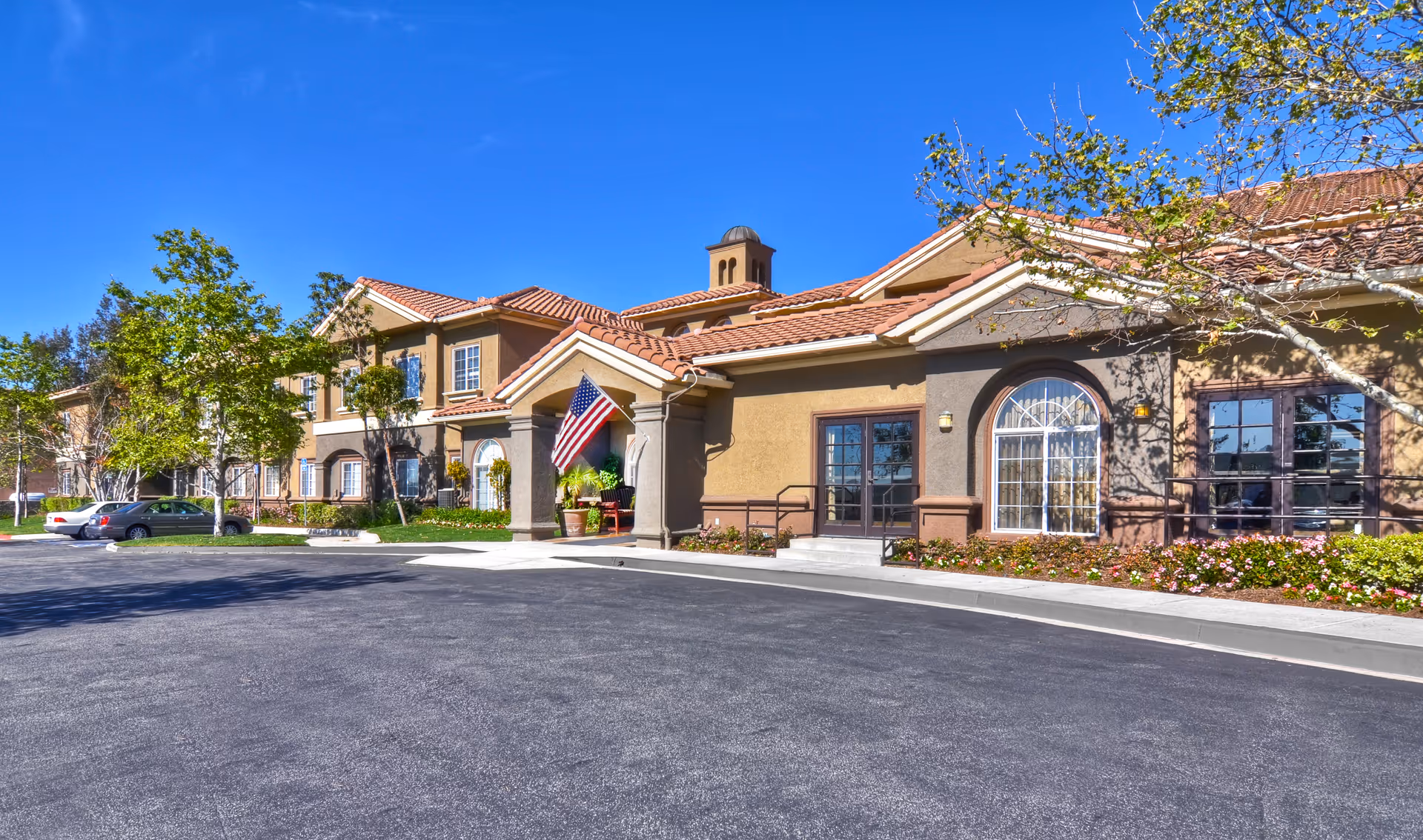 Exterior view of a senior living facility building with a beige and brown facade, red tile roof, and an American flag displayed near the entrance. There are trees, shrubs, and flowers around the building, and a paved driveway in front under a clear blue sky.