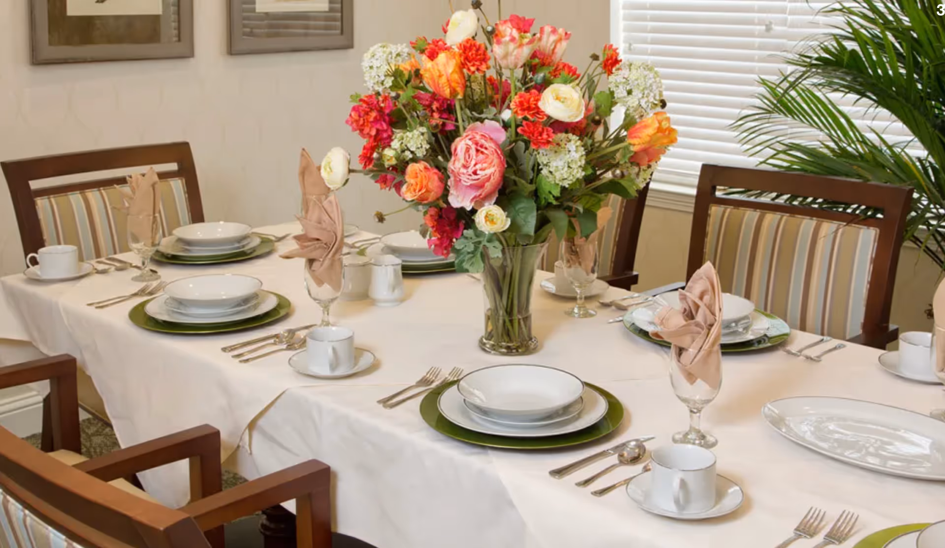 A formally set dining table with plates, folded napkins, silverware, and a large floral centerpiece in a dining room.