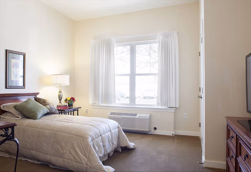 A bright bedroom with a single bed covered in a beige comforter and green pillows. There is a wooden nightstand with a lamp, books, and a small vase of flowers next to the bed. A large window with white curtains lets in natural light, and an air conditioning unit is installed below the window. A wooden dresser with a TV on top is visible on the right side of the room.