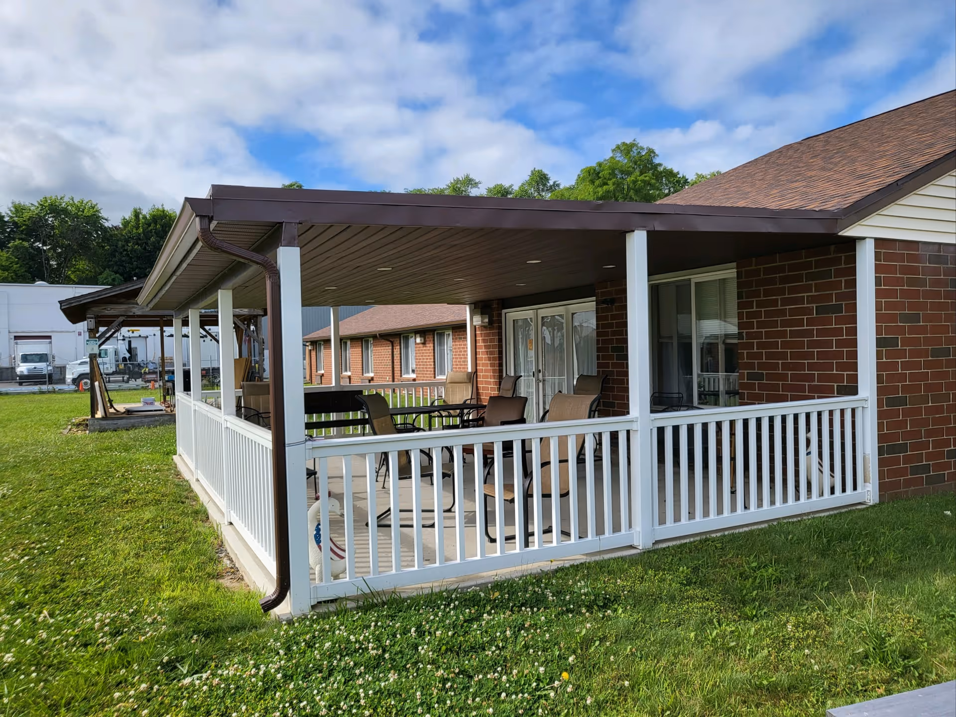 Covered outdoor patio with white railing and seating attached to a brick building under a partly cloudy sky.
