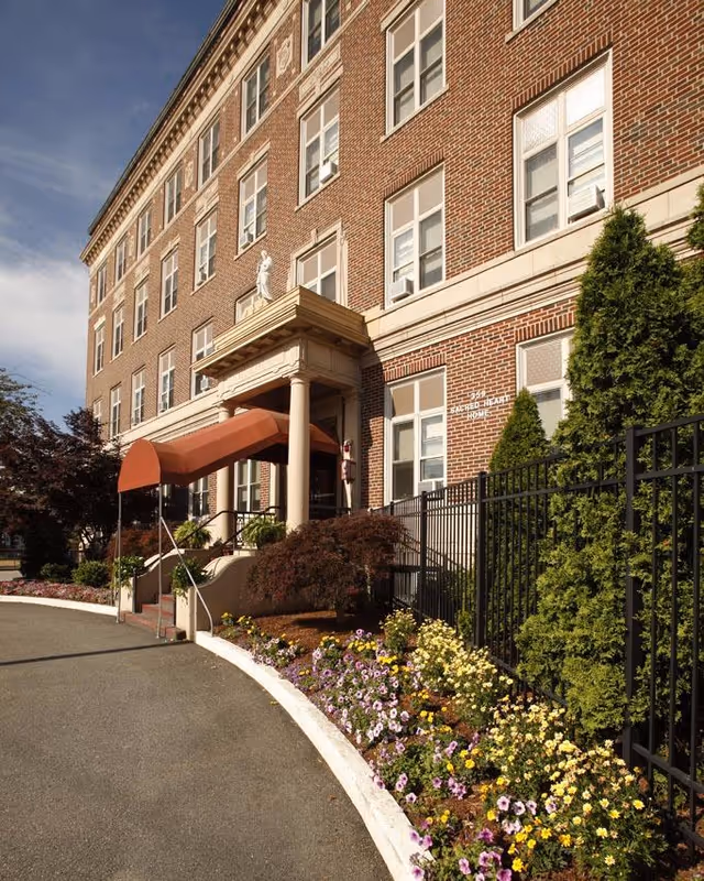 Front exterior of a brick senior living building with a covered entrance, columns, and flower beds along a curved driveway.