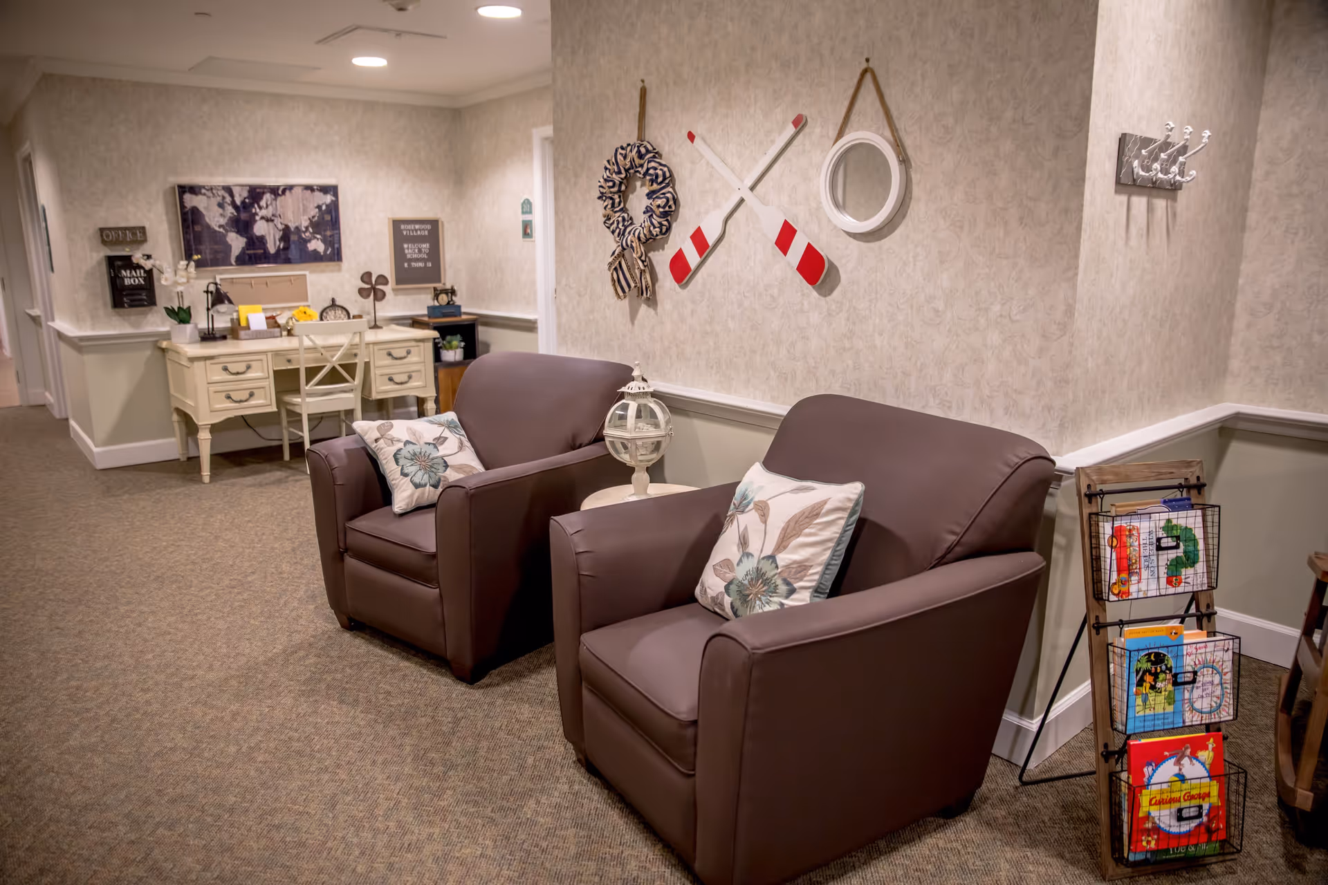 A cozy interior corner of RoseWood Village Assisted Living & Memory Care - Greenbrier featuring two brown armchairs with floral cushions, a small round table with a decorative lantern between them, and a wall decorated with a wreath, two crossed oars, and a round mirror. In the background, there is a cream-colored desk with a chair, a world map on the wall, and various decorative items including a mailbox and a sign. A wooden rack holding colorful books is positioned near the armchairs.