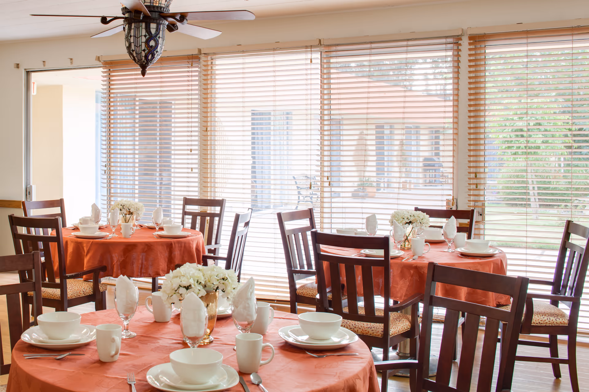 Dining area with round tables covered in orange tablecloths, set with white plates, bowls, cups, and napkins. Each table has a centerpiece of white flowers. Wooden chairs surround the tables, and large windows with wooden blinds let in natural light, showing an outdoor view.