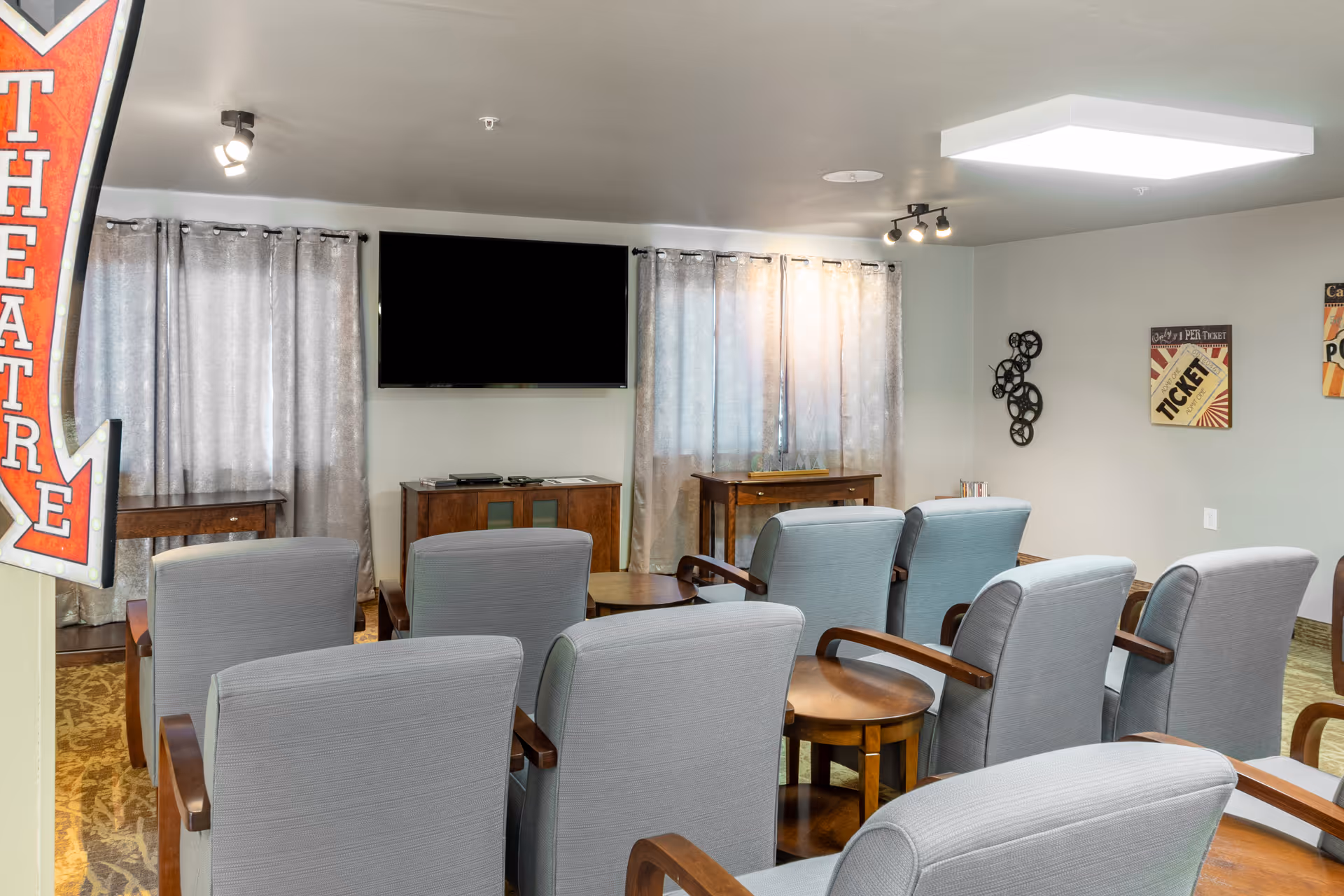 Small theater-style seating area with rows of armchairs facing a wall-mounted TV in a senior living common room.