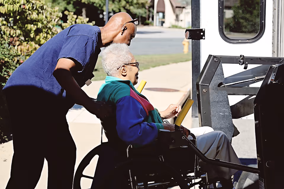 A caregiver assists an elderly woman in a wheelchair as they approach a vehicle equipped with a wheelchair lift outdoors on a sunny day.