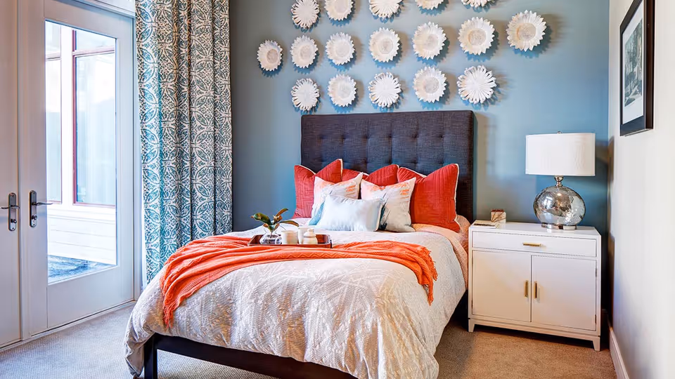 A cozy bedroom with a dark upholstered headboard, a bed dressed in white and coral bedding with multiple pillows, a white nightstand with a silver lamp, decorative white wall art above the bed, patterned curtains, and glass doors leading outside.