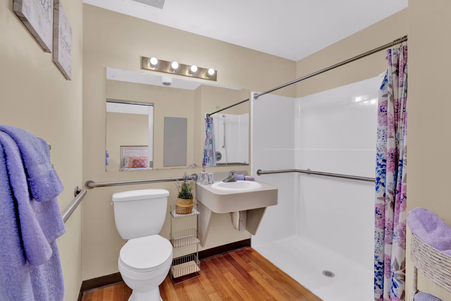 A clean and accessible bathroom with beige walls and wood flooring. It features a white toilet, a wall-mounted sink with a small shelf underneath, and a large mirror above the sink. There are grab bars around the toilet and inside the walk-in shower area, which has a floral shower curtain in shades of purple, blue, and pink. Purple towels hang on a towel rack and a basket with purple towels is visible on the right side. The bathroom is well-lit with a four-bulb light fixture above the mirror.