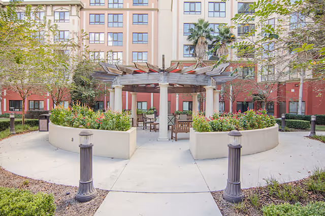 Outdoor courtyard area with a wooden pergola supported by white columns, surrounded by raised flower beds and seating. The courtyard is part of a multi-story building with windows and red and beige exterior walls. Trees and landscaped plants are visible around the paved walkway.