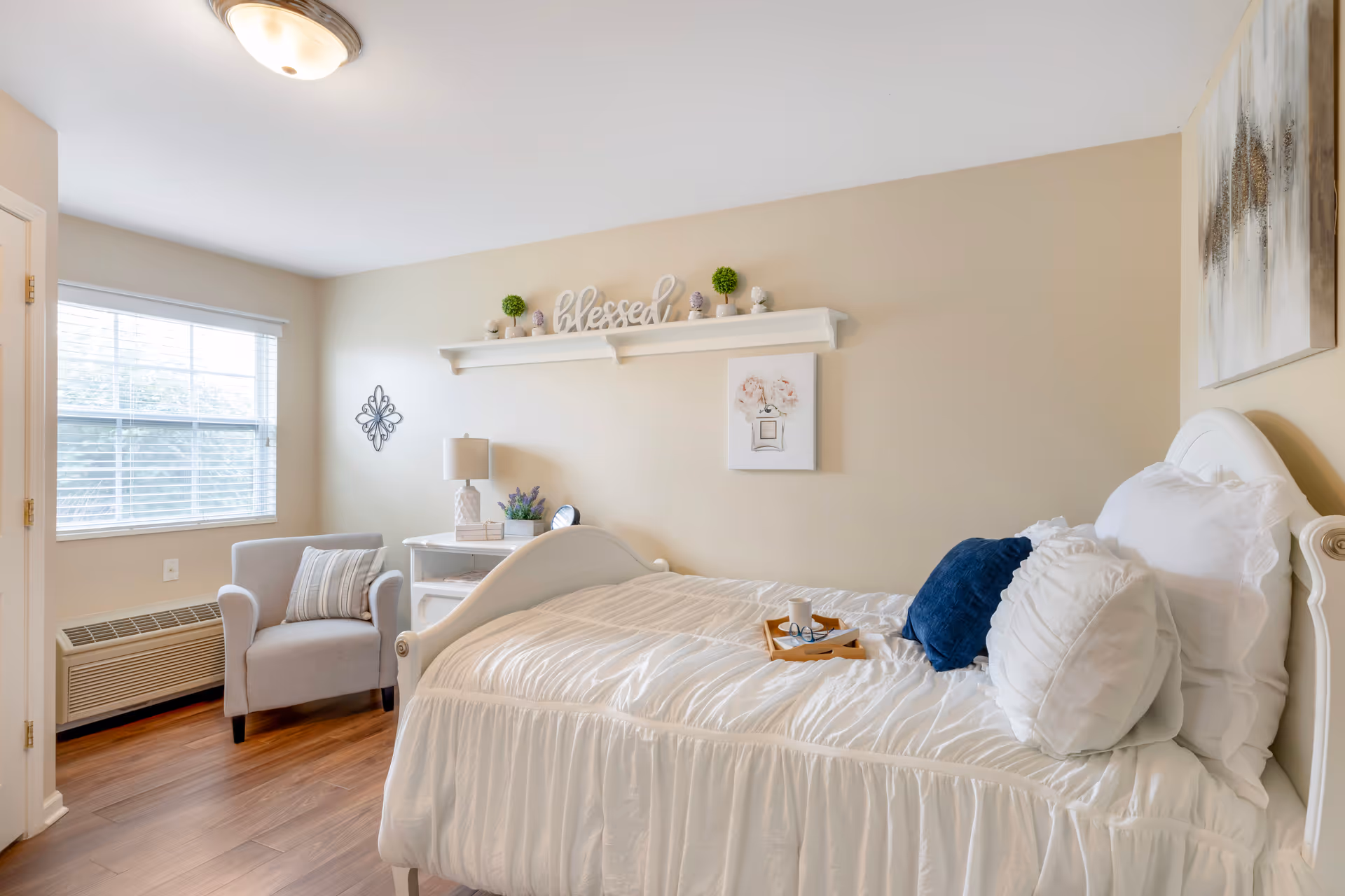 A bright bedroom with a white bed dressed in white linens and pillows, including a blue accent pillow. A wooden tray with a cup and glasses rests on the bed. There is a gray armchair with a striped pillow near a window with blinds. A white desk with a lamp, books, and a small plant is against the wall. Above the desk is a shelf with decorative items and the word 'blessed'. The walls are light beige, and there is a framed floral picture and abstract artwork on the walls. The floor is wooden.
