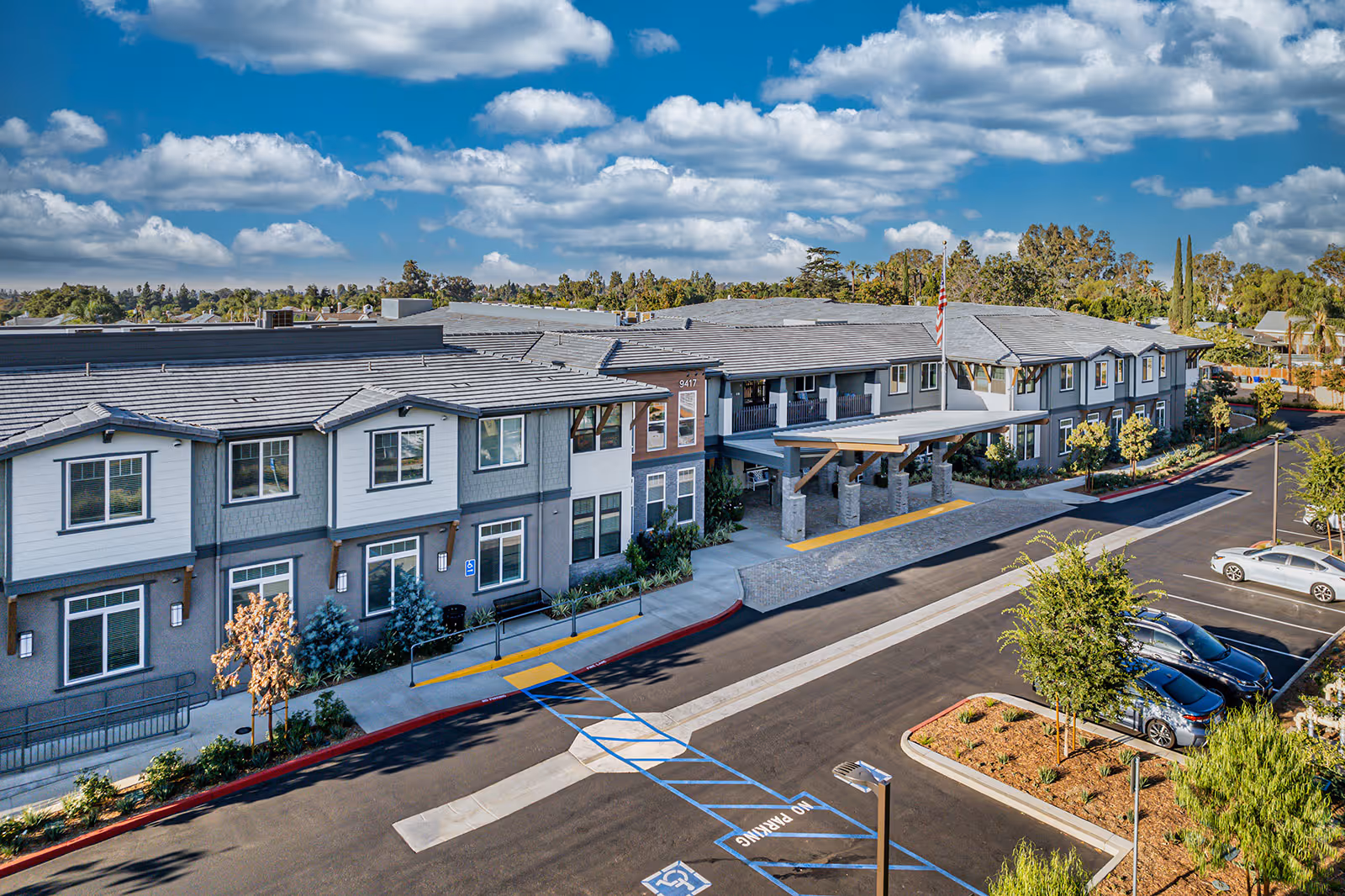 Exterior view of Allara Senior Living facility showing a modern two-story building with a covered entrance, parking spaces including handicapped spots, landscaped areas with trees and shrubs, under a partly cloudy blue sky.
