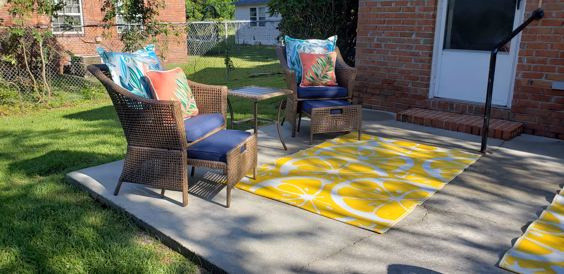 Outdoor patio area with two wicker chairs featuring blue cushions and colorful leaf-patterned pillows, a small wicker footrest, a small table between the chairs, and a yellow rug with white lemon slice patterns on a concrete surface next to a brick building with a white door and black handrail.