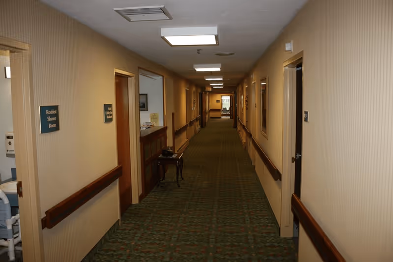 A long indoor hallway in a senior living facility with beige walls, green patterned carpet, and wooden handrails on both sides. Several doors line the hallway, with signs indicating a Resident Shower Room and Staff Utility and Linen Room on the left. The hallway is lit by ceiling lights and has a small table with items on it along the left wall.
