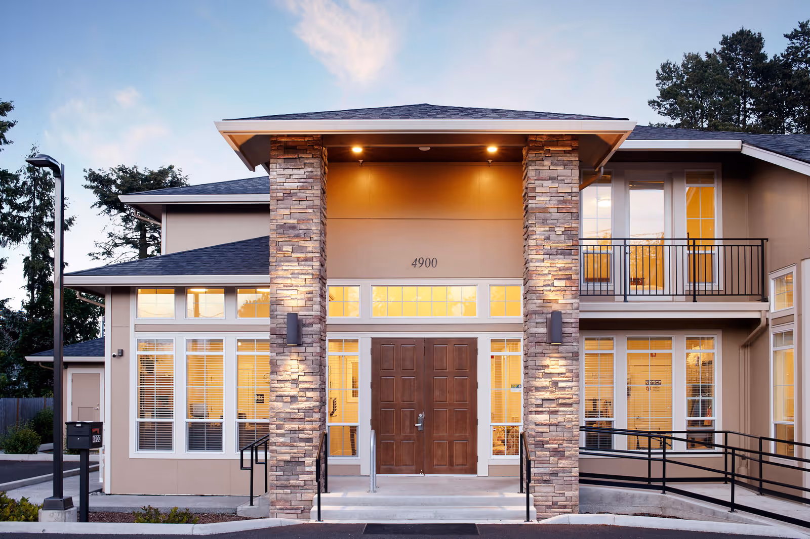 Front entrance of a two-story memory care building with stone pillars, large windows, a balcony, and double wooden doors lit from inside.