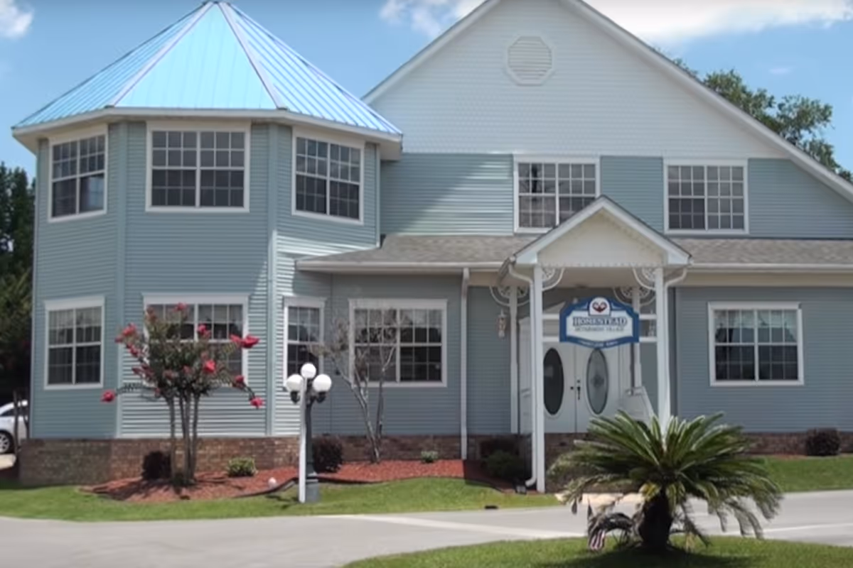 Exterior view of a two-story building with light blue siding and a blue metal roof. The building has multiple windows and a small covered entrance with a sign that reads 'Homestead of Pensacola'. There are landscaped areas with small trees, bushes, and a lamp post in front of the building under a clear blue sky.