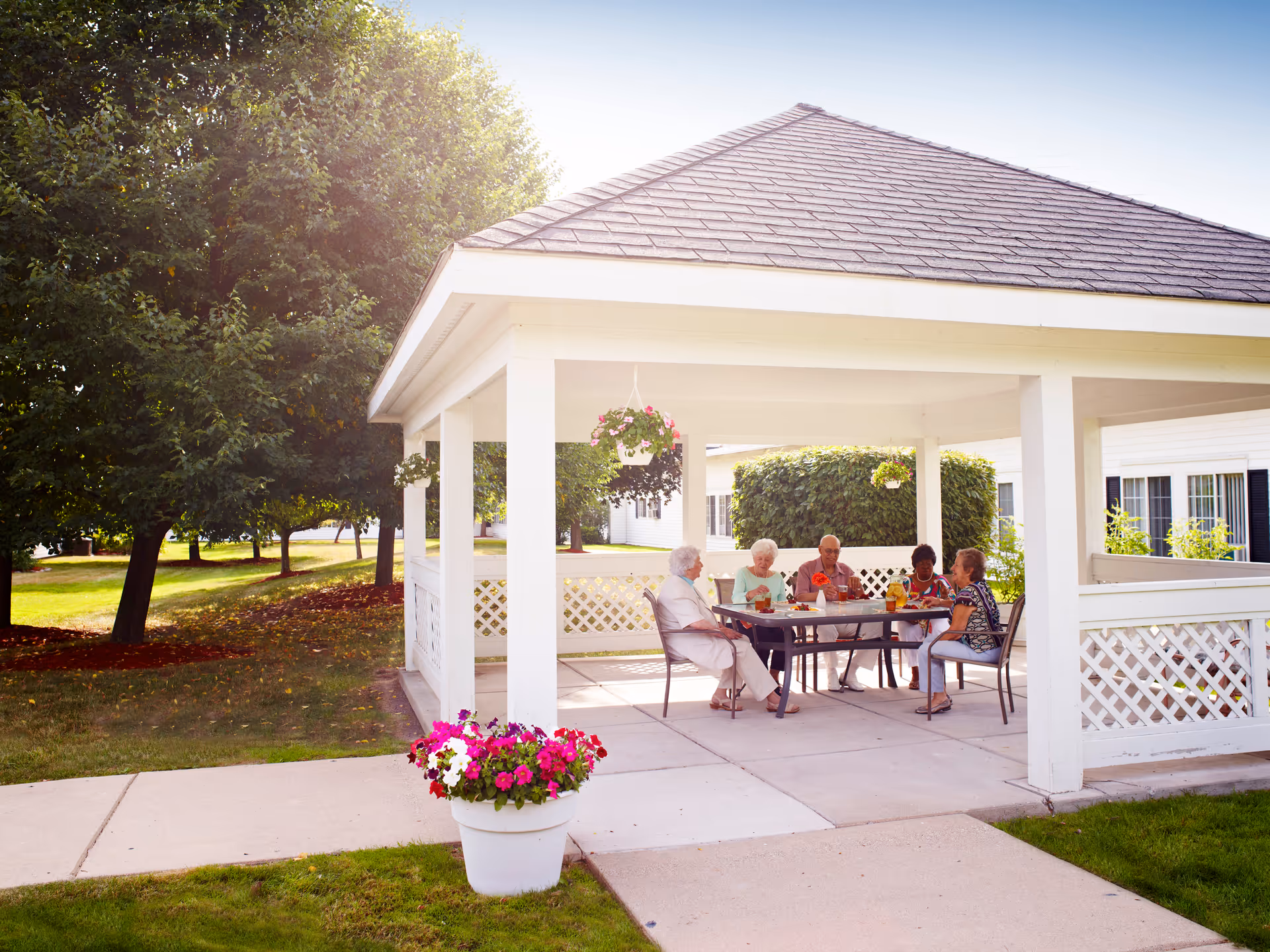 A group of elderly people sitting around a table under a white gazebo in a garden area, enjoying drinks and conversation. The gazebo is surrounded by greenery, including trees and bushes, with hanging flower pots and a flower pot on the ground nearby.