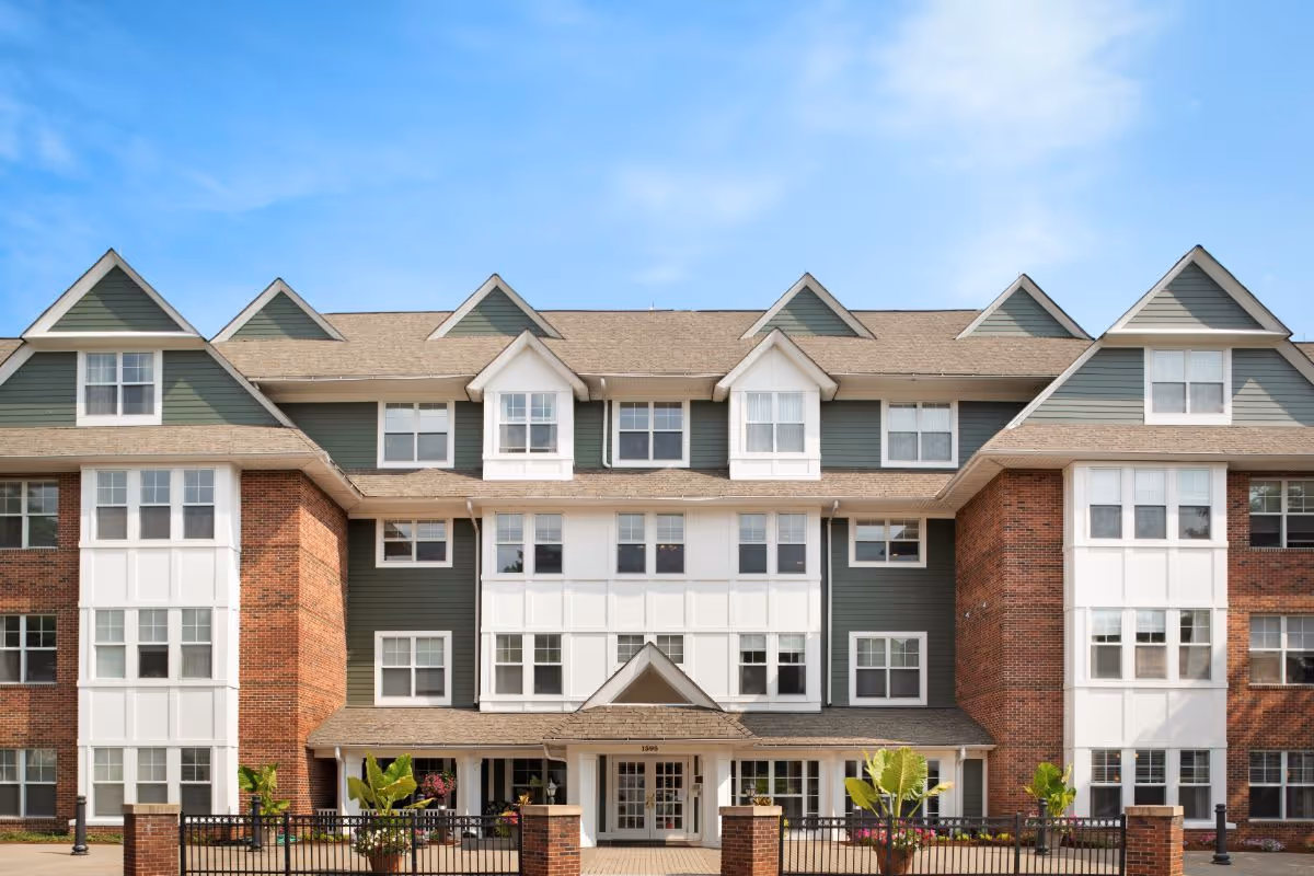 Front exterior view of a multi-story senior living facility building with green and white siding, brick accents, multiple windows, and a central entrance with a small covered porch. There are plants and flowers near the entrance and a black metal fence in front.