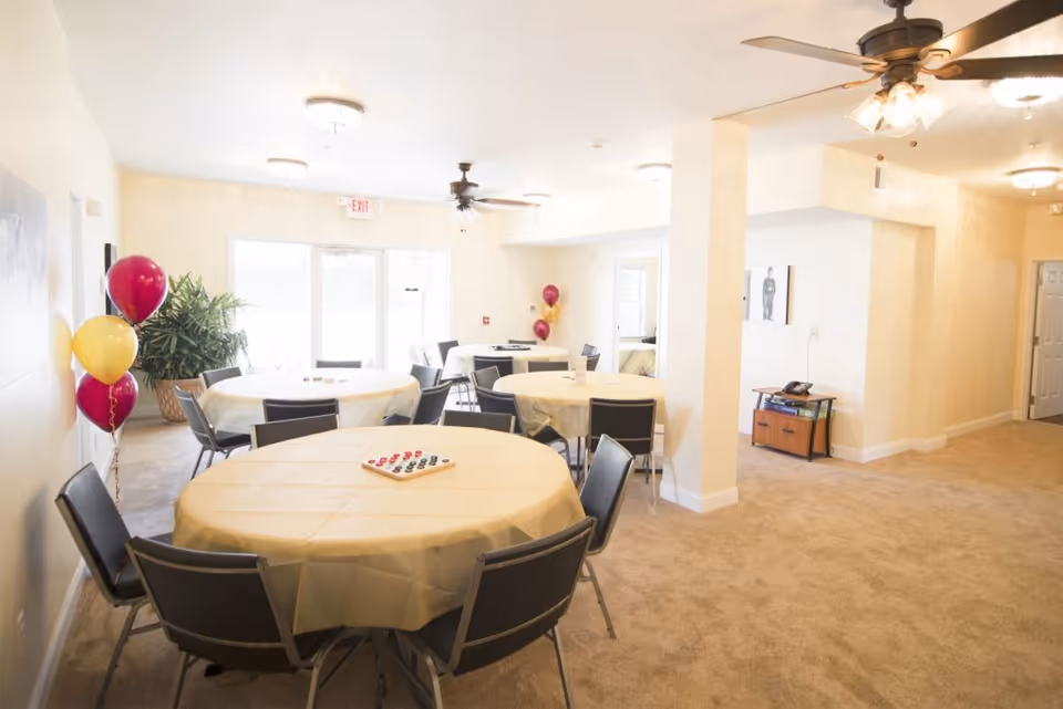 A bright, carpeted room with round tables covered in beige tablecloths and surrounded by black chairs. There are ceiling fans with lights, a few balloons in red and yellow colors, a potted plant near the glass exit doors, and a small table with a phone and some items against the wall. The room appears to be a communal or activity space in a senior living facility.