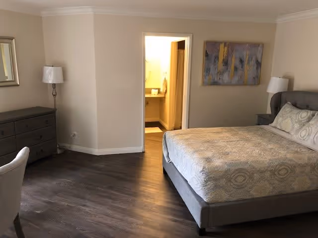 A bedroom with a large bed featuring patterned bedding, a nightstand with a lamp, a dresser with a lamp, a chair, and an open door leading to a bathroom with a sink and mirror visible.