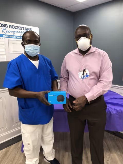 Two men standing indoors in a room with gray walls and wood flooring. One man is wearing blue scrubs and a face mask, holding a boxed Echo Dot device. The other man is wearing a pink shirt, dark pants, an N95 mask, and an RN badge. Behind them is a purple-covered table and a wall sign that reads 'ROSS ROCKSTARS Recognize'.