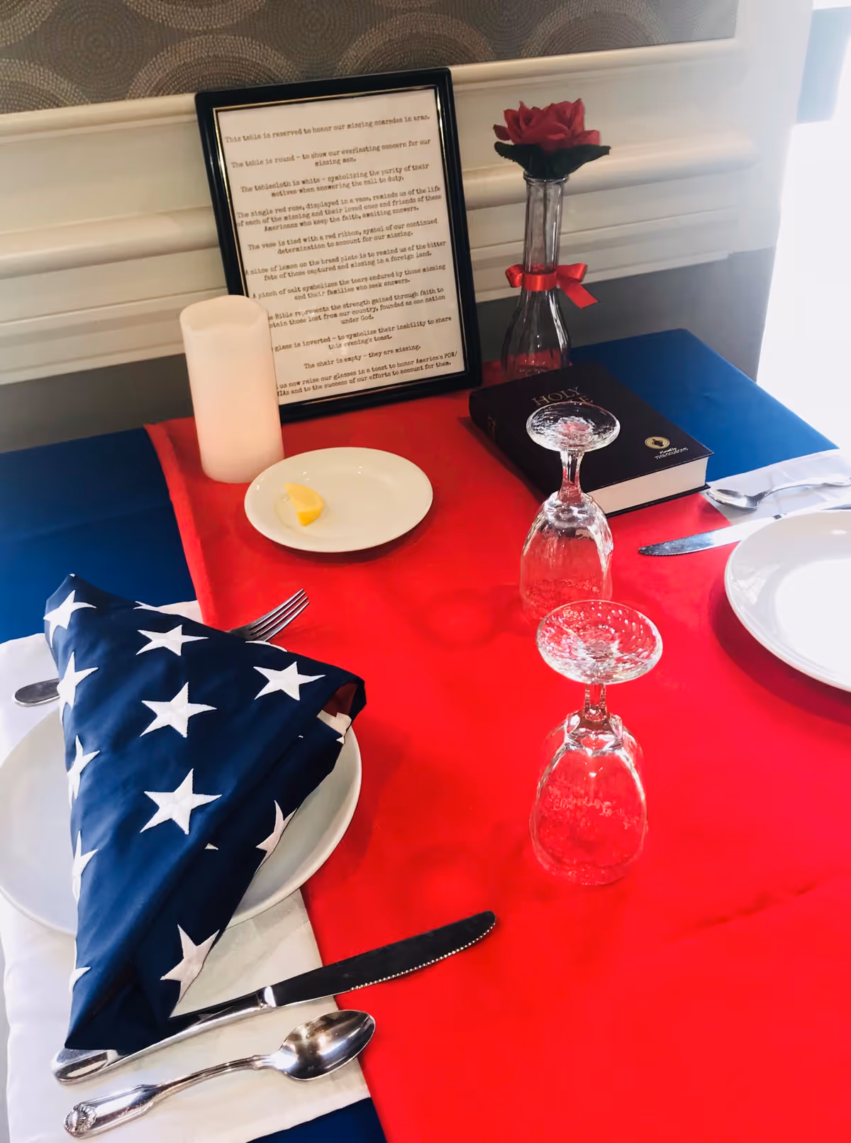 A dining table set with a red tablecloth and a folded American flag napkin on a white plate. There are utensils including a knife, fork, and spoon placed around the plate. Two upside-down wine glasses are on the table, along with a white plate holding a lemon wedge. A framed poem or text, a lit candle, a red rose in a glass vase with a red ribbon, and a Holy Bible are also on the table.