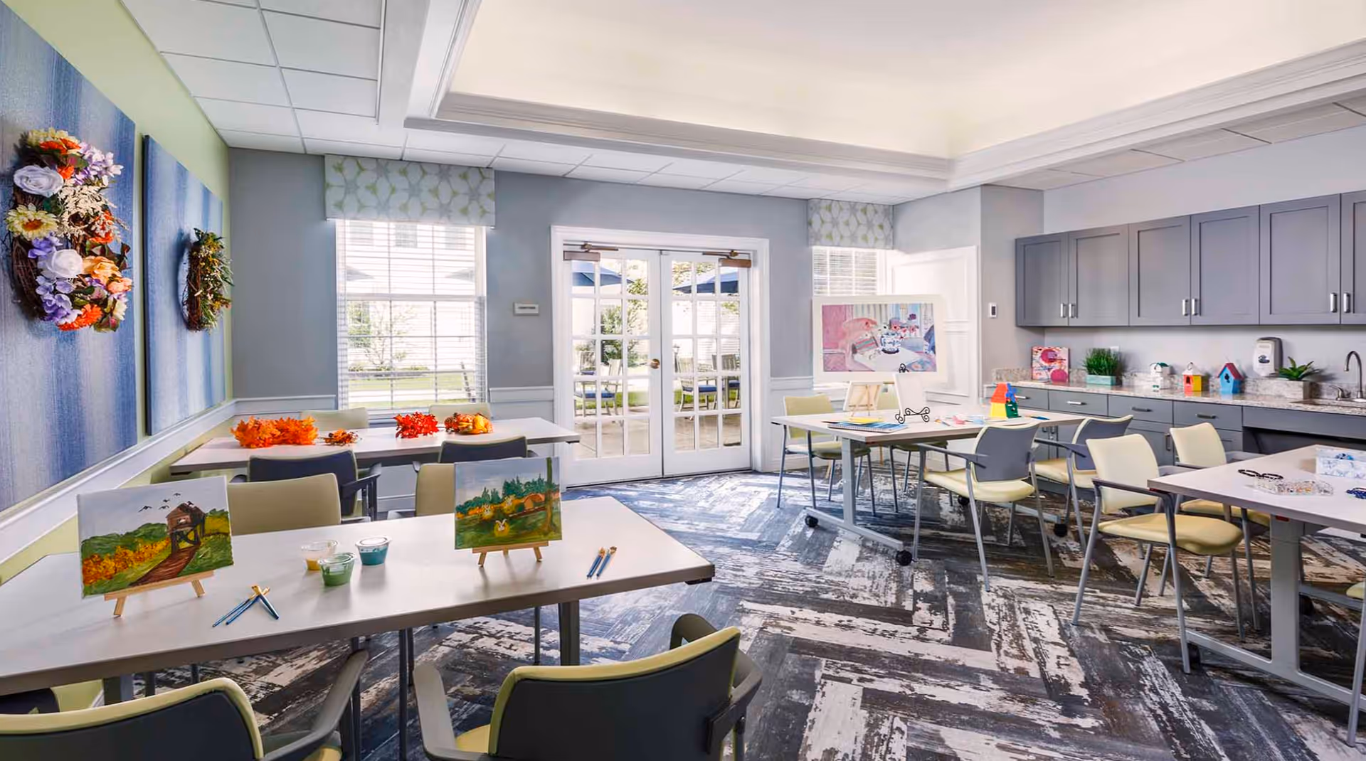 A bright and spacious activity room with several tables and chairs arranged for arts and crafts. Paintings on small easels, paintbrushes, and paint cups are placed on the tables. The room has large windows and glass doors leading outside, with light blue and green walls decorated with floral wreaths and patterned valances. Gray cabinets and a countertop with various supplies are visible along one wall.