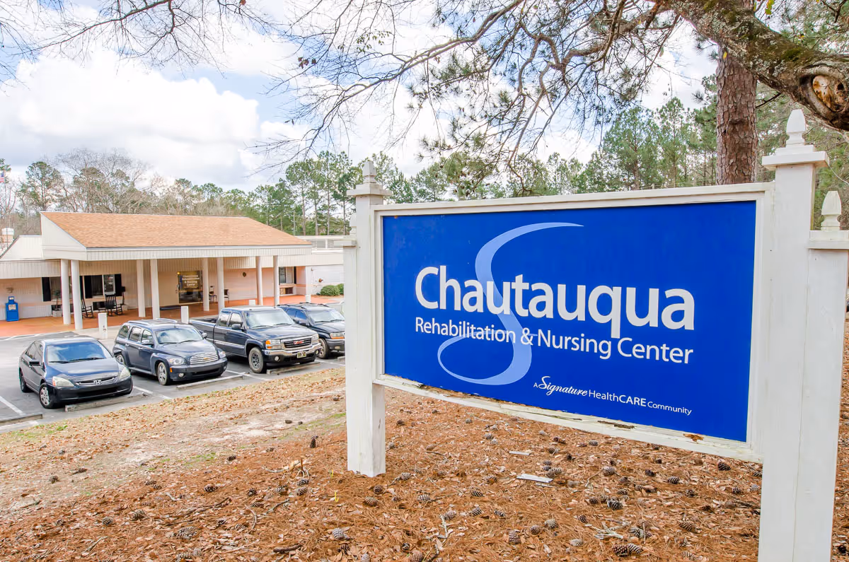 Exterior view of Chautauqua Rehabilitation & Nursing Center with a blue sign in the foreground and a parking lot with several cars in front of the building. Trees and a partly cloudy sky are visible in the background.
