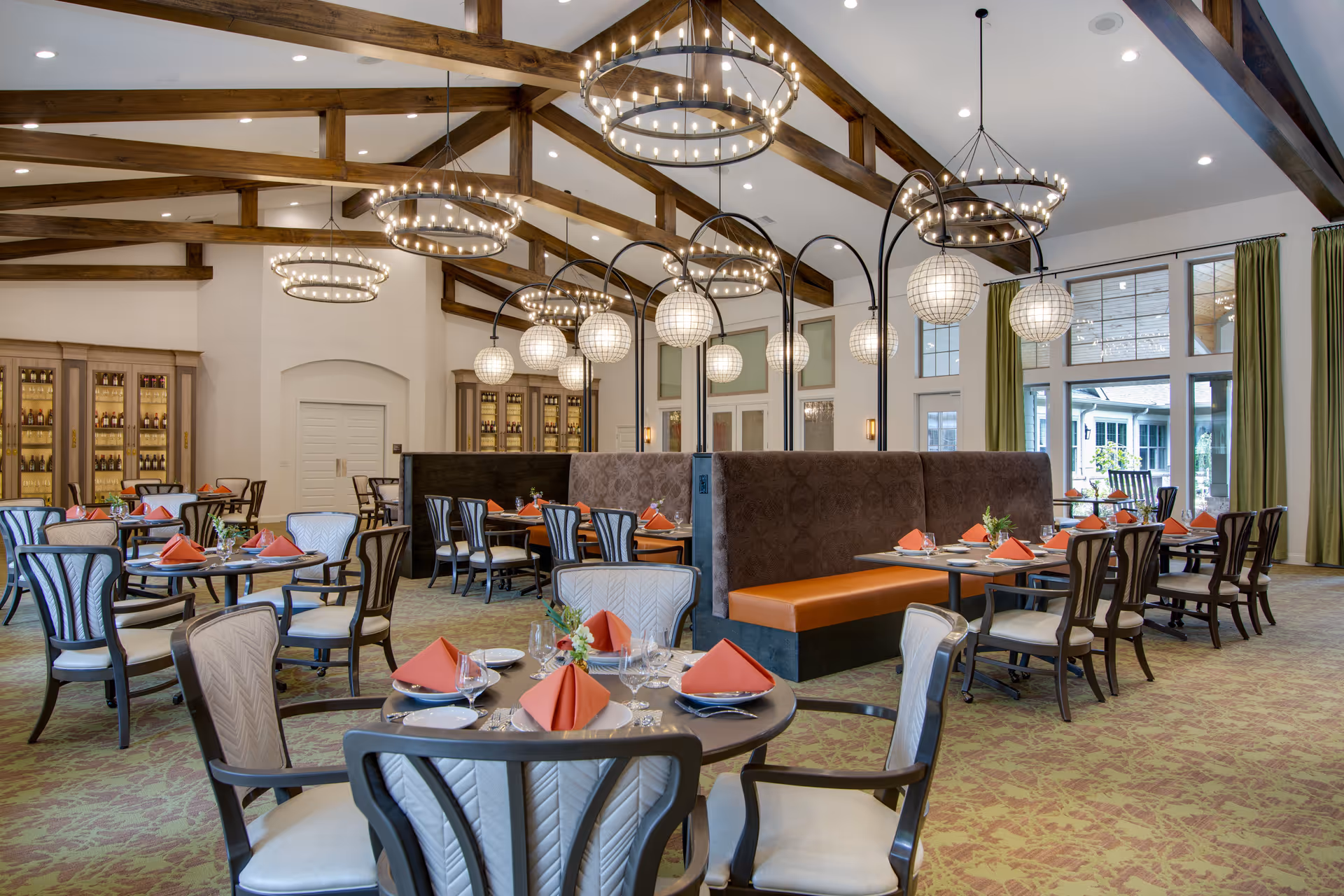 Spacious dining room with round and rectangular tables set with plates, glasses, and orange folded napkins. The room features high vaulted ceilings with exposed wooden beams and multiple circular chandeliers. There are large windows with green curtains allowing natural light to enter, and cushioned booth seating with globe-shaped hanging lamps above.