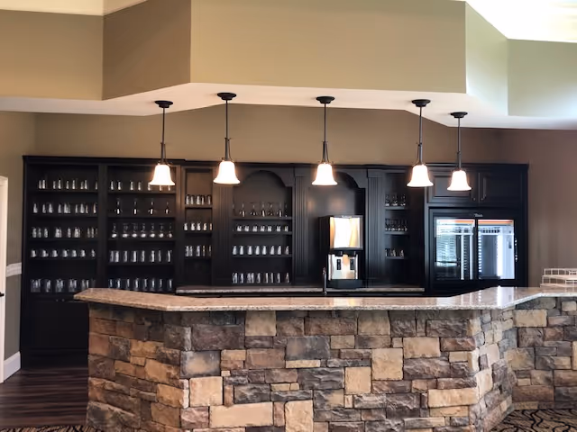 Interior view of a bar area with a stone facade counter and dark wooden shelves filled with glassware. Five pendant lights hang from the ceiling above the counter. There is a beverage dispenser and a glass-front refrigerator behind the counter.