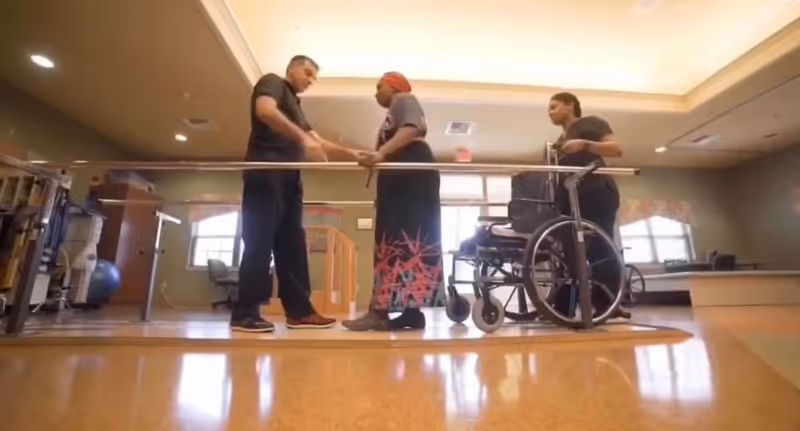 A healthcare worker assists an elderly woman standing and holding hands in a rehabilitation or therapy room. Another person stands nearby holding a wheelchair. The room has parallel bars for physical therapy and exercise equipment in the background.