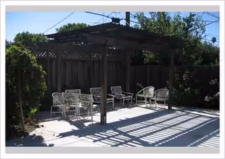 Outdoor patio with metal chairs under a wooden pergola, surrounded by fencing.