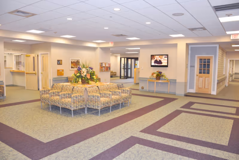 A spacious and well-lit common area in an assisted living facility with patterned carpet flooring, a circular arrangement of cushioned chairs around a table with a floral centerpiece, a wall-mounted television, and multiple doorways leading to other rooms and hallways.