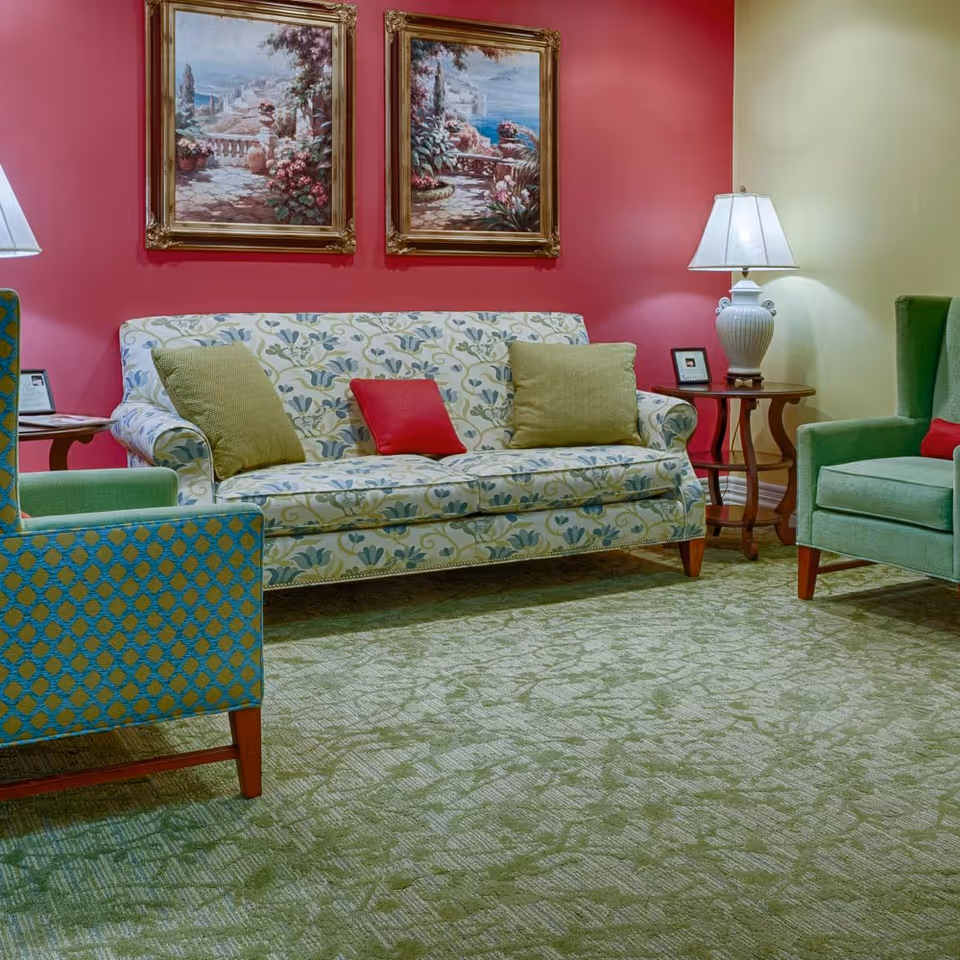 A cozy living room area with a floral patterned sofa adorned with green and red cushions, two green armchairs, a green patterned carpet, two framed landscape paintings on a red accent wall, and two white table lamps on wooden side tables.