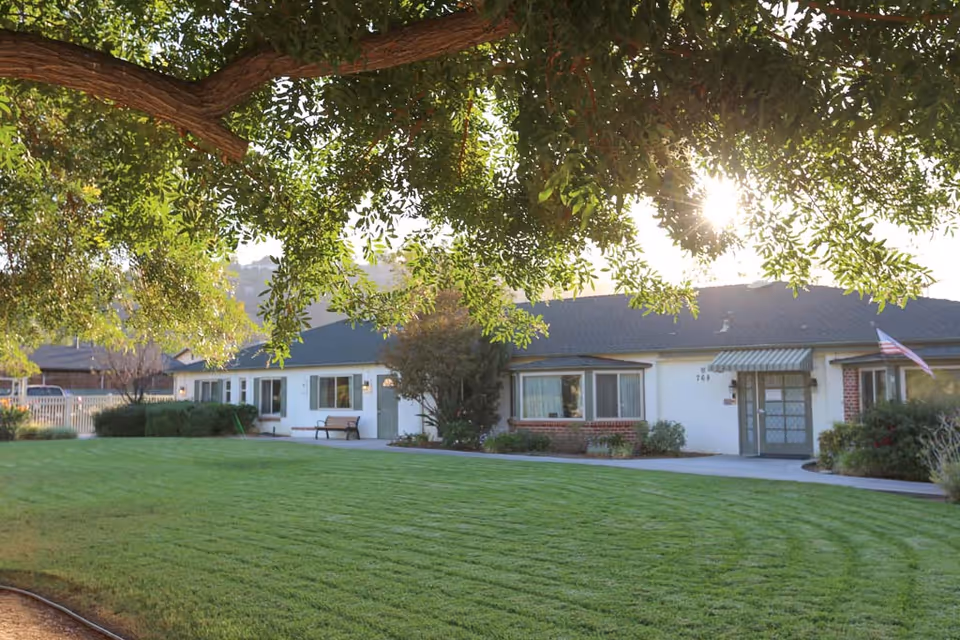 A single-story senior living facility building with a well-maintained green lawn in front, surrounded by trees and shrubs. The sun is shining through the tree branches above, casting light on the building and lawn. There is a bench near the entrance and an American flag displayed near the door.
