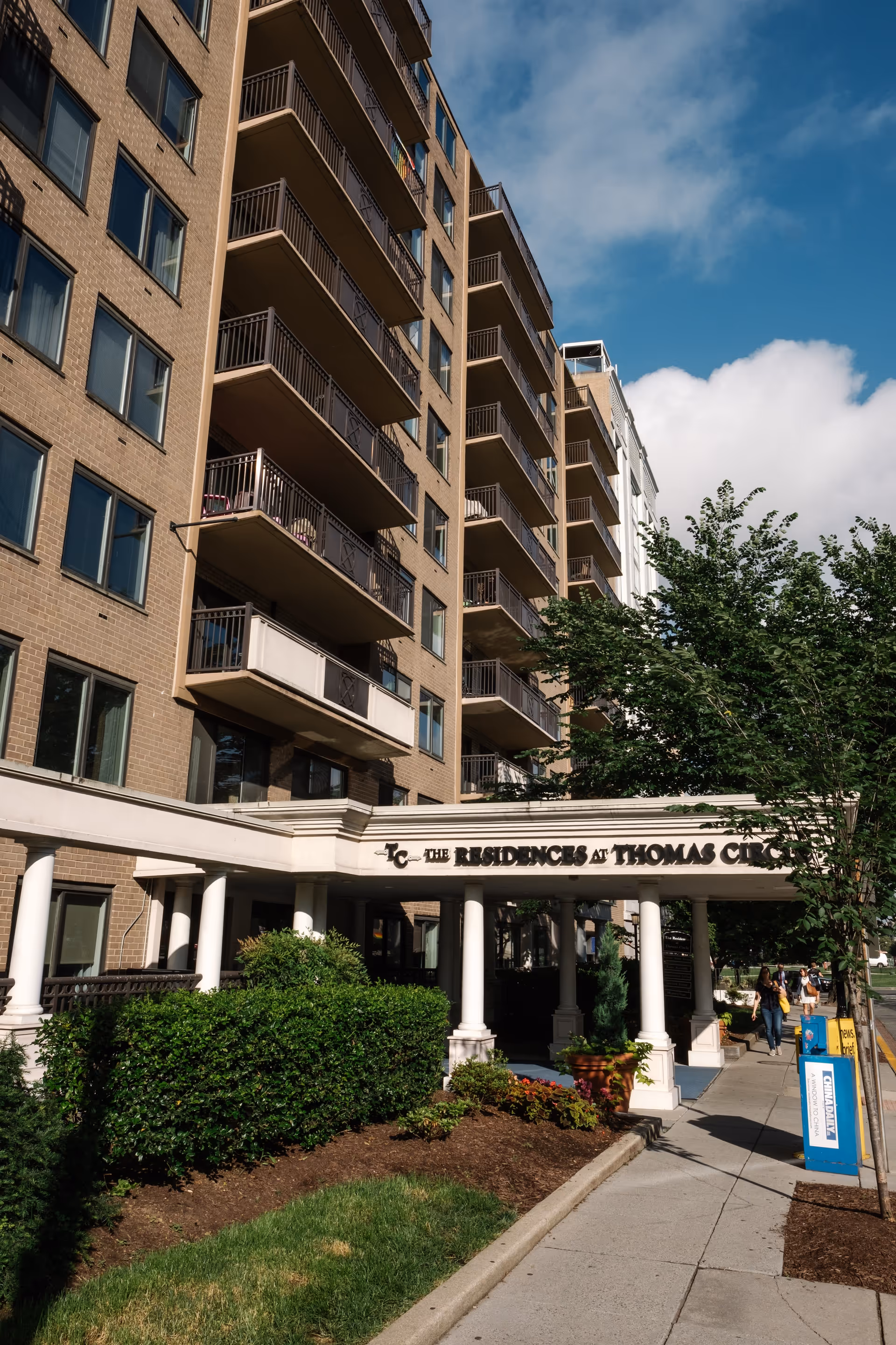 Exterior view of a multi-story residential building with balconies and a covered entrance labeled 'The Residences at Thomas Circle.' There are green bushes and trees near the entrance, and a sidewalk with pedestrians walking by.