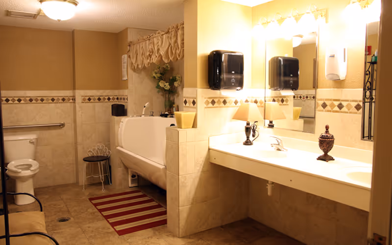 A well-lit bathroom with beige tiled walls and floor, featuring a toilet with a grab bar, a large bathtub with decorative flowers and a curtain valance above, a double sink vanity with a large mirror, two black soap dispensers, a small lamp, and decorative items on the counter.