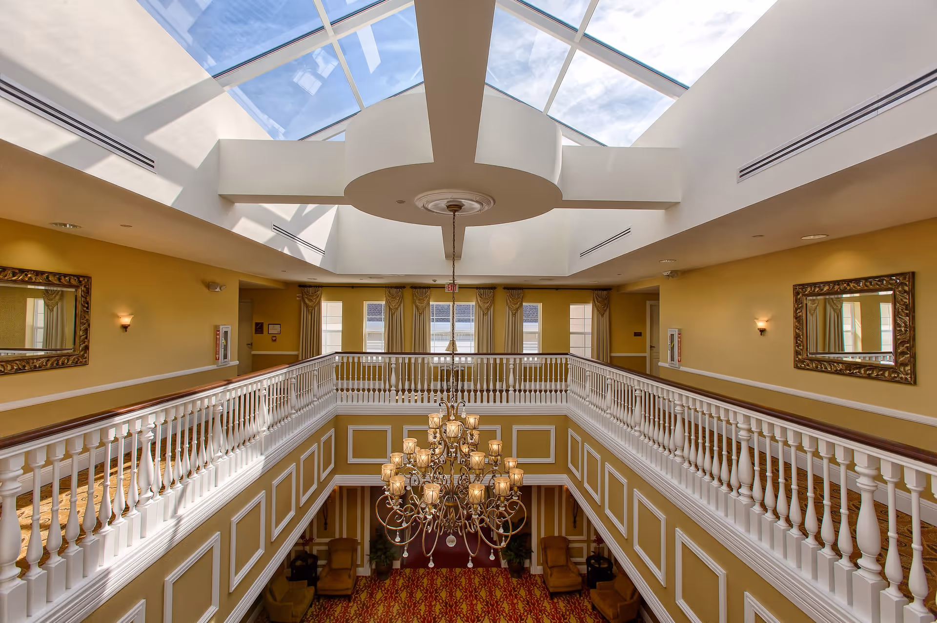 Bright two-story interior atrium with a large skylight, central chandelier, and white balustrade balcony overlooking a seating area.