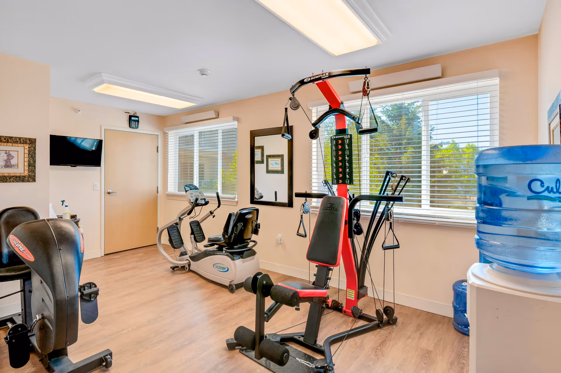 A small fitness room with exercise equipment including a Bowflex machine, a recumbent exercise bike, and another cardio machine. The room has light-colored walls, wood flooring, two large windows with white blinds letting in natural light, a wall-mounted TV, a mirror, framed pictures, and a water cooler with a large blue water jug.