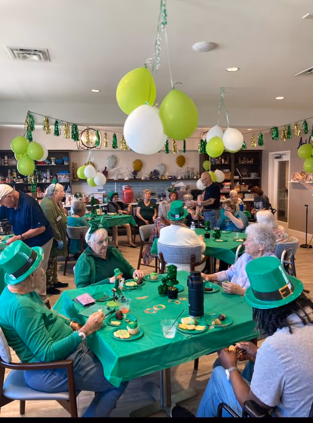 Seniors gathered in a decorated communal dining room wearing green and seated at tables with green tablecloths and balloons for a celebration.