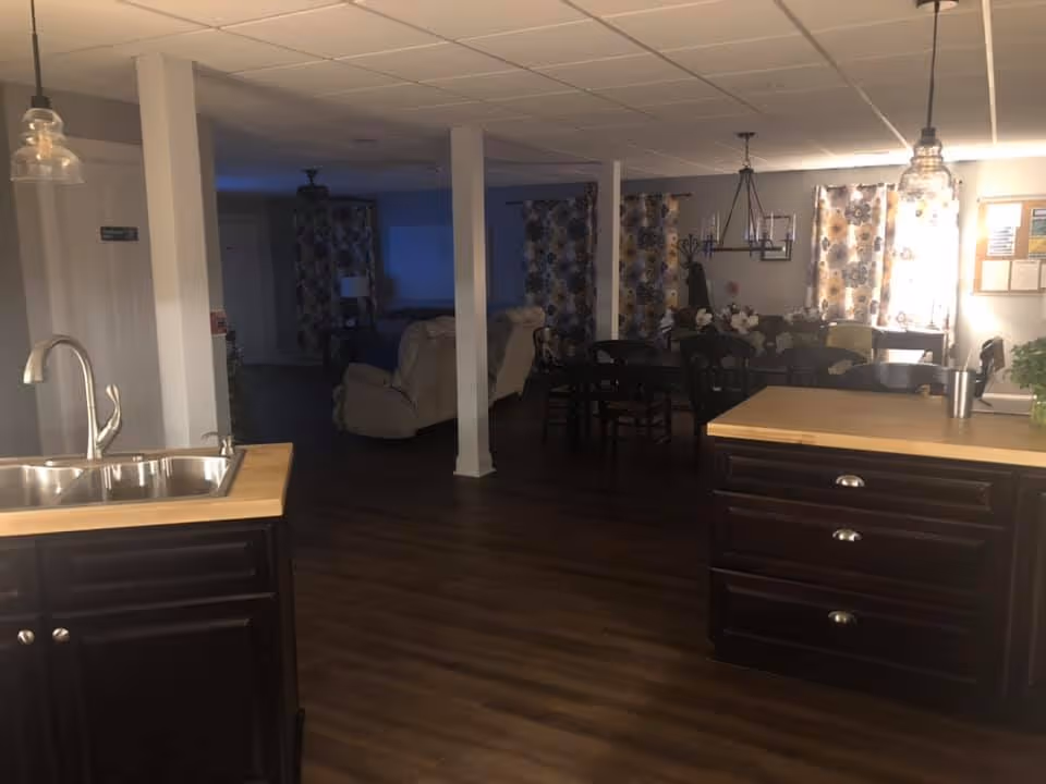 Interior view of a living space in Magnolia Assisted Living showing a kitchen area with a sink and wooden countertops in the foreground, and a dining area with a table and chairs in the background. The room has dark wood flooring, white ceiling tiles, and floral curtains on the windows. There are also some comfortable recliner chairs and a chandelier above the dining table.