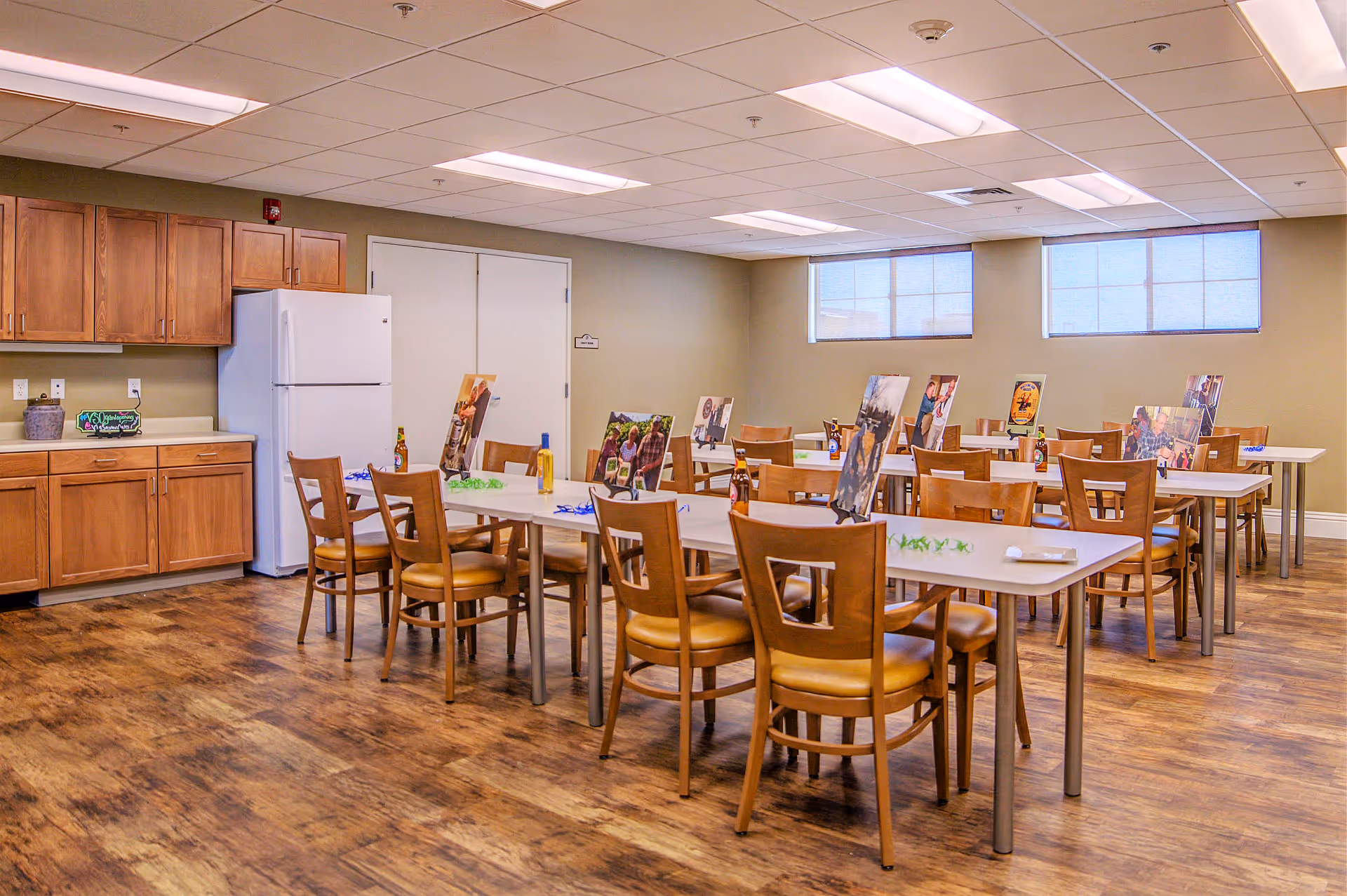 A well-lit dining room with wooden chairs and tables arranged in rows. Each table has bottles and photo displays on stands. The room has wooden cabinets, a white refrigerator, and two windows with blinds. The floor is wooden, and the ceiling has fluorescent lights.