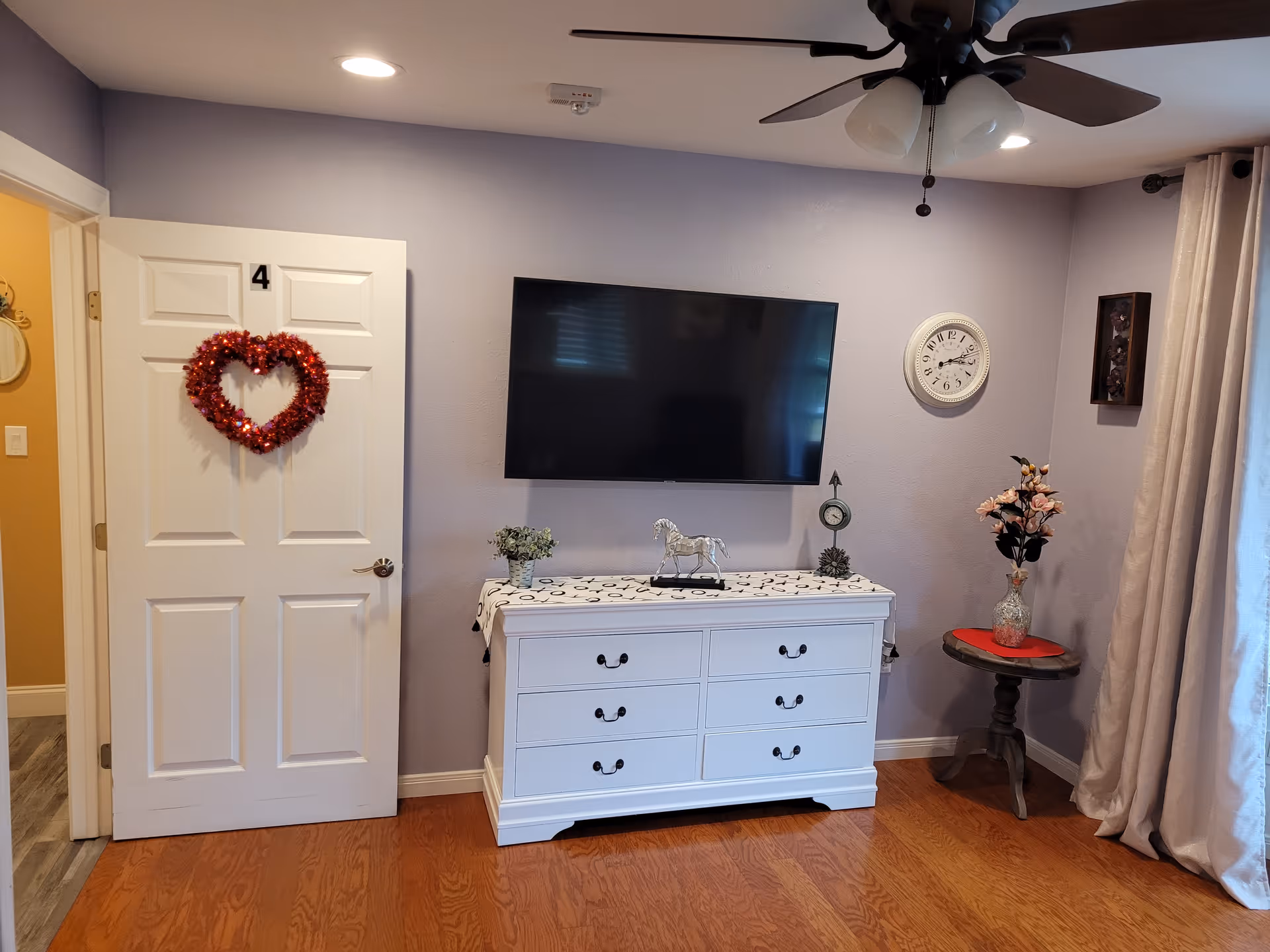 A cozy room with a wall-mounted TV above a white dresser, a door labeled '4' with a red heart wreath, a small side table with flowers, and a ceiling fan.