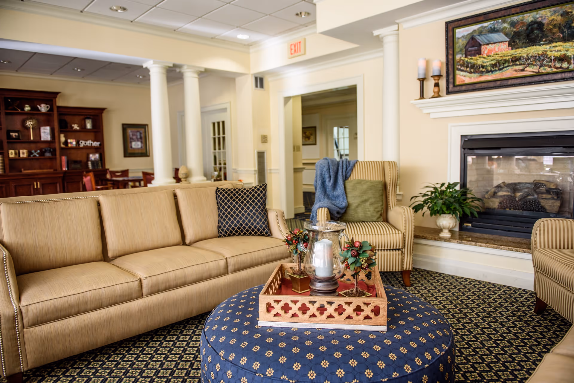 A cozy living room area with a beige sofa and striped armchairs surrounding a round blue ottoman with a decorative tray holding candles and small holiday decorations. A fireplace with a painting above it and a potted plant on the hearth is visible on the right. In the background, there is a wooden cabinet with shelves and a dining area with tables and chairs. The room has cream-colored walls, white columns, and patterned carpet.