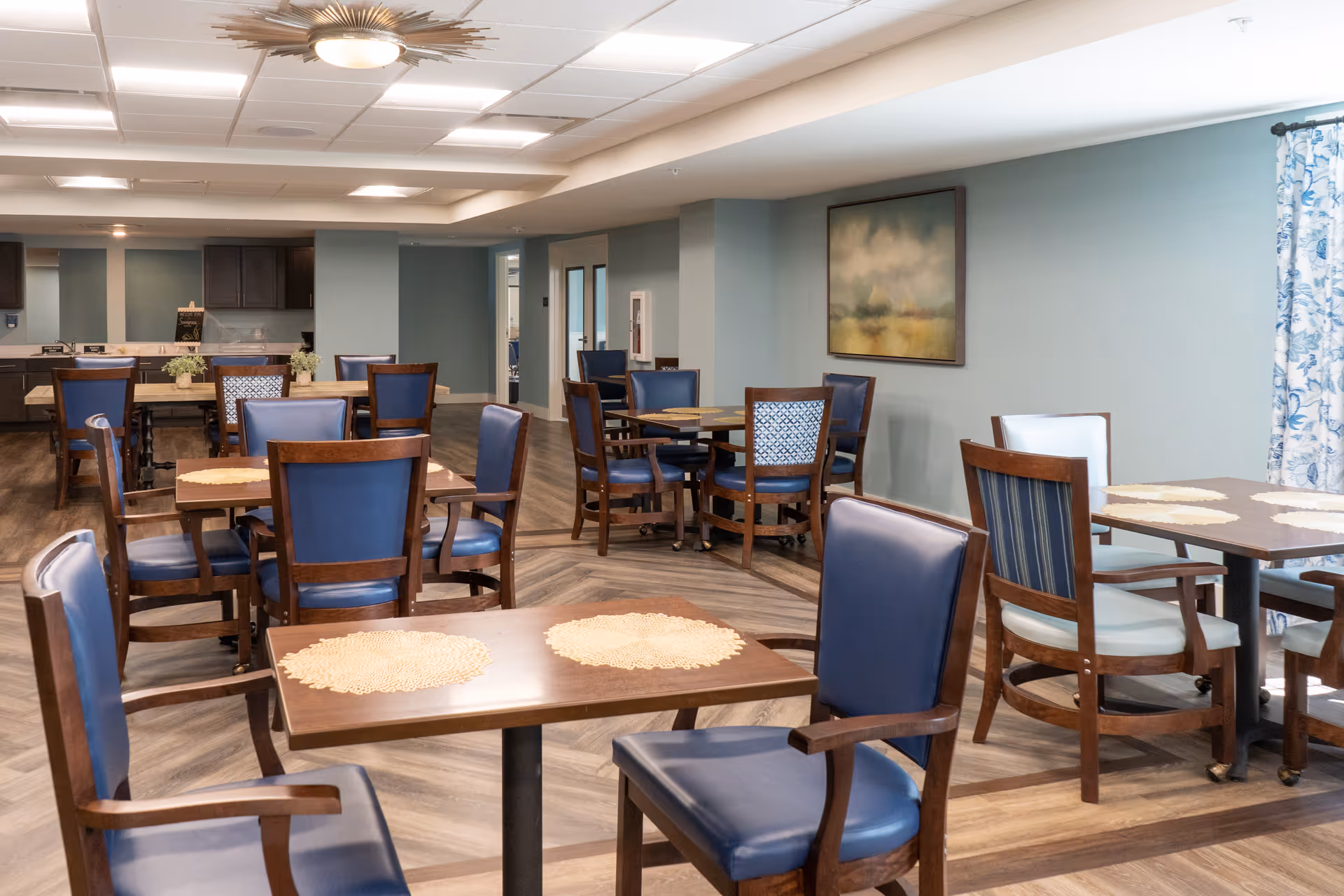 A dining room with several wooden tables and chairs with blue and white cushions. The room has light blue walls, a large painting, patterned curtains, and a modern ceiling light fixture. The floor has a wood pattern, and there is a kitchen area visible in the background.