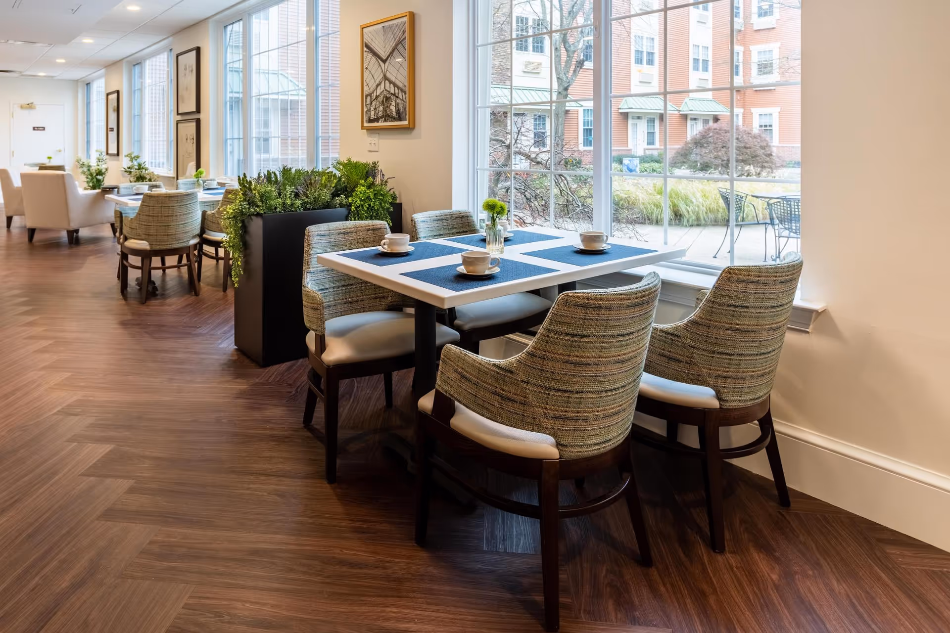 A bright dining area in a senior living facility with several tables and cushioned chairs. The tables are set with placemats, cups, and small flower vases. Large windows allow natural light to fill the room and offer a view of an outdoor garden area with shrubs and patio furniture. The floor has a wood pattern, and framed artwork decorates the walls.