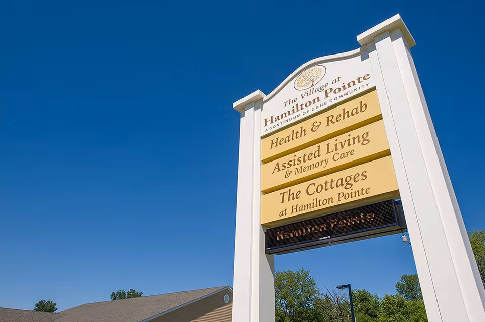 Outdoor image of a tall sign for The Village at Hamilton Pointe against a clear blue sky. The sign lists services including Health & Rehab, Assisted Living & Memory Care, and The Cottages at Hamilton Pointe. Trees and part of a building roof are visible in the background.