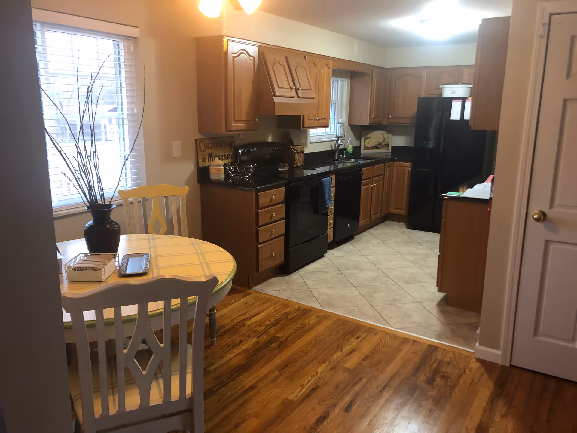 Small open kitchen with wooden cabinets, black appliances, tiled floor, and a nearby round dining table on hardwood flooring.