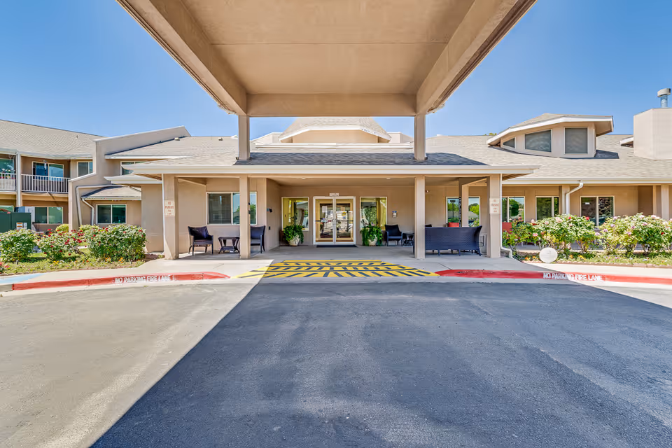 Front entrance and covered drop-off area of a senior living facility with seating, planters, and a marked fire lane.