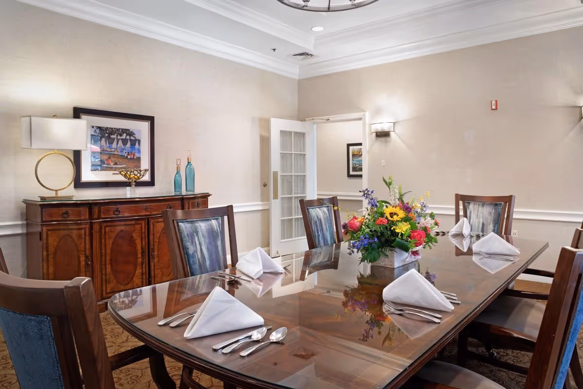 Formal dining room with a polished wooden table set with folded napkins, silverware and a floral centerpiece.