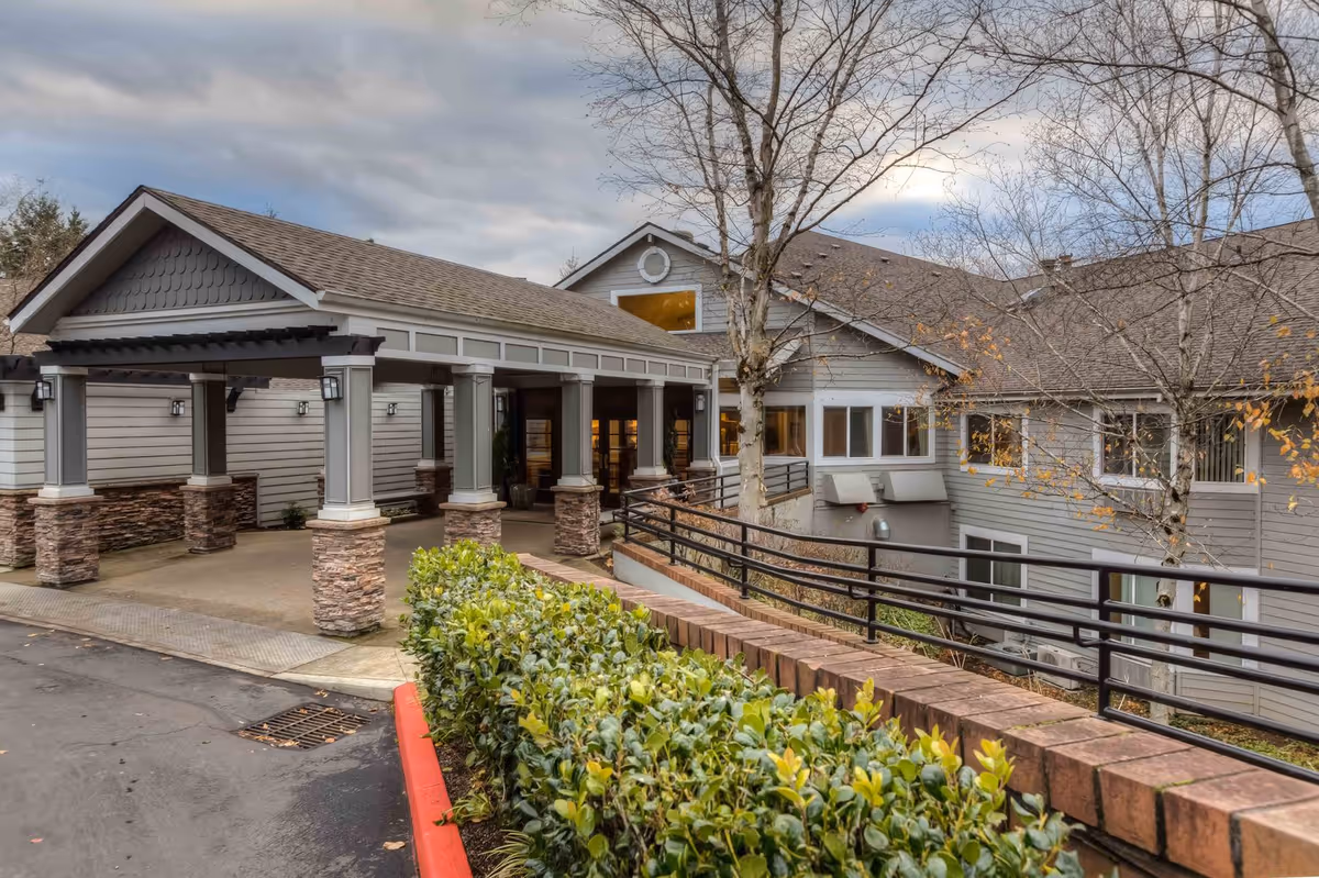 Covered entrance and porte-cochere of a multi-story senior living building with stone-column supports, ramped railings, and landscaping.